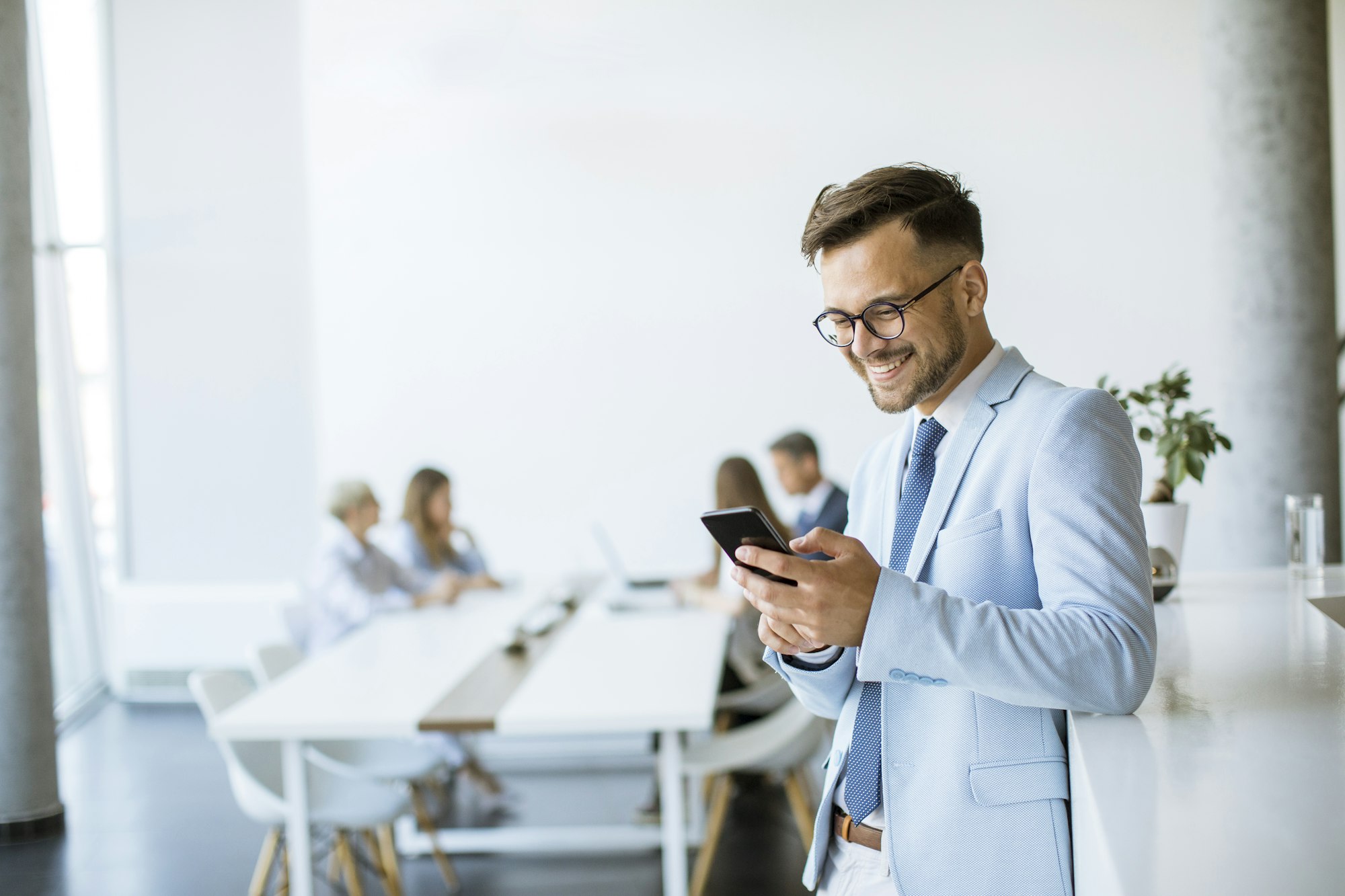 Happy young man using his mobile phone and smiling while his colleagues working in the background Happy young man using his mobile phone and smiling while his colleagues working in the background
